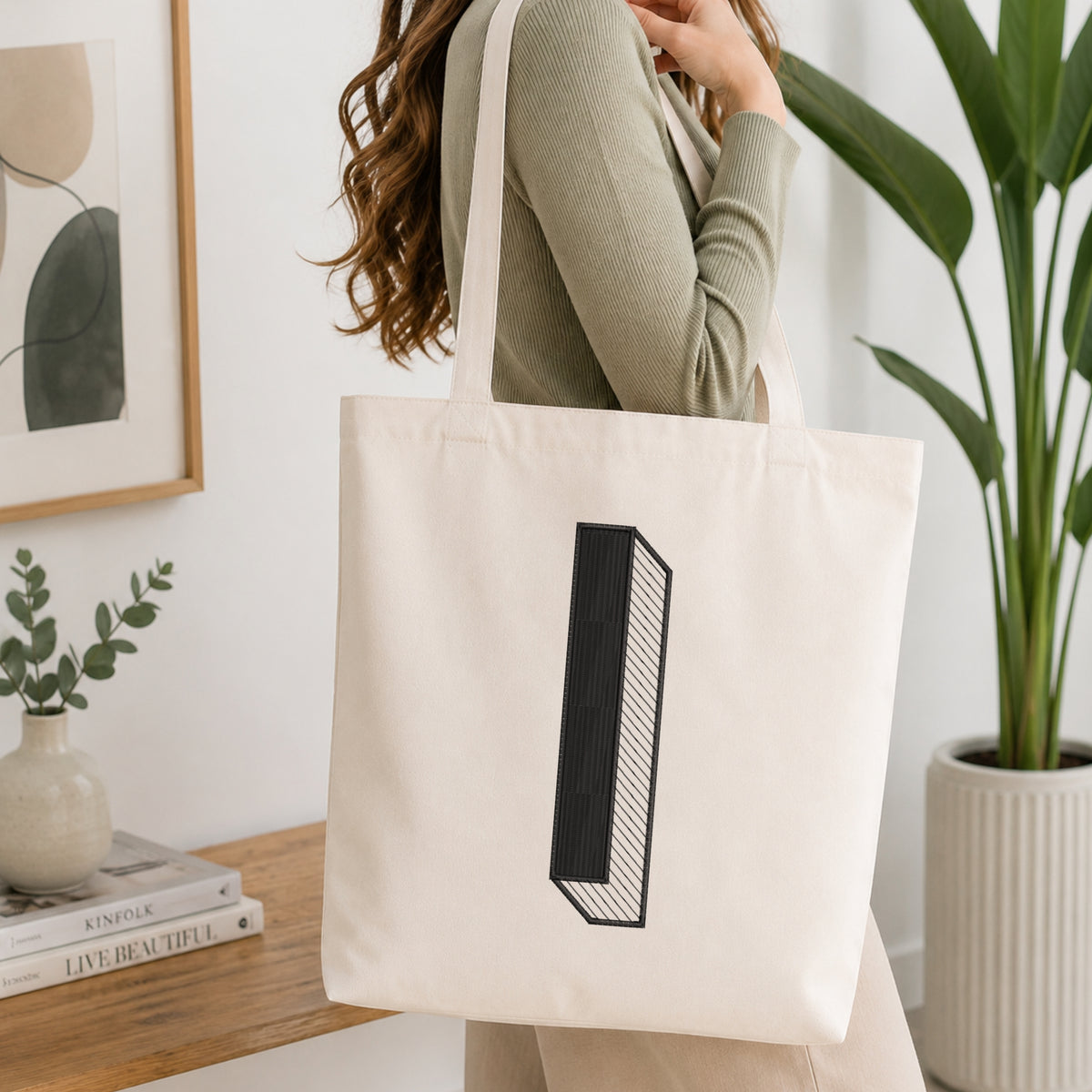 A woman wearing a sage green ribbed long-sleeve top carries a large off-white canvas tote bag with a black geometric block graphic on the front. The scene is set in a bright, modern room with a wooden console table, a framed print, a small vase with greenery, and a potted plant.