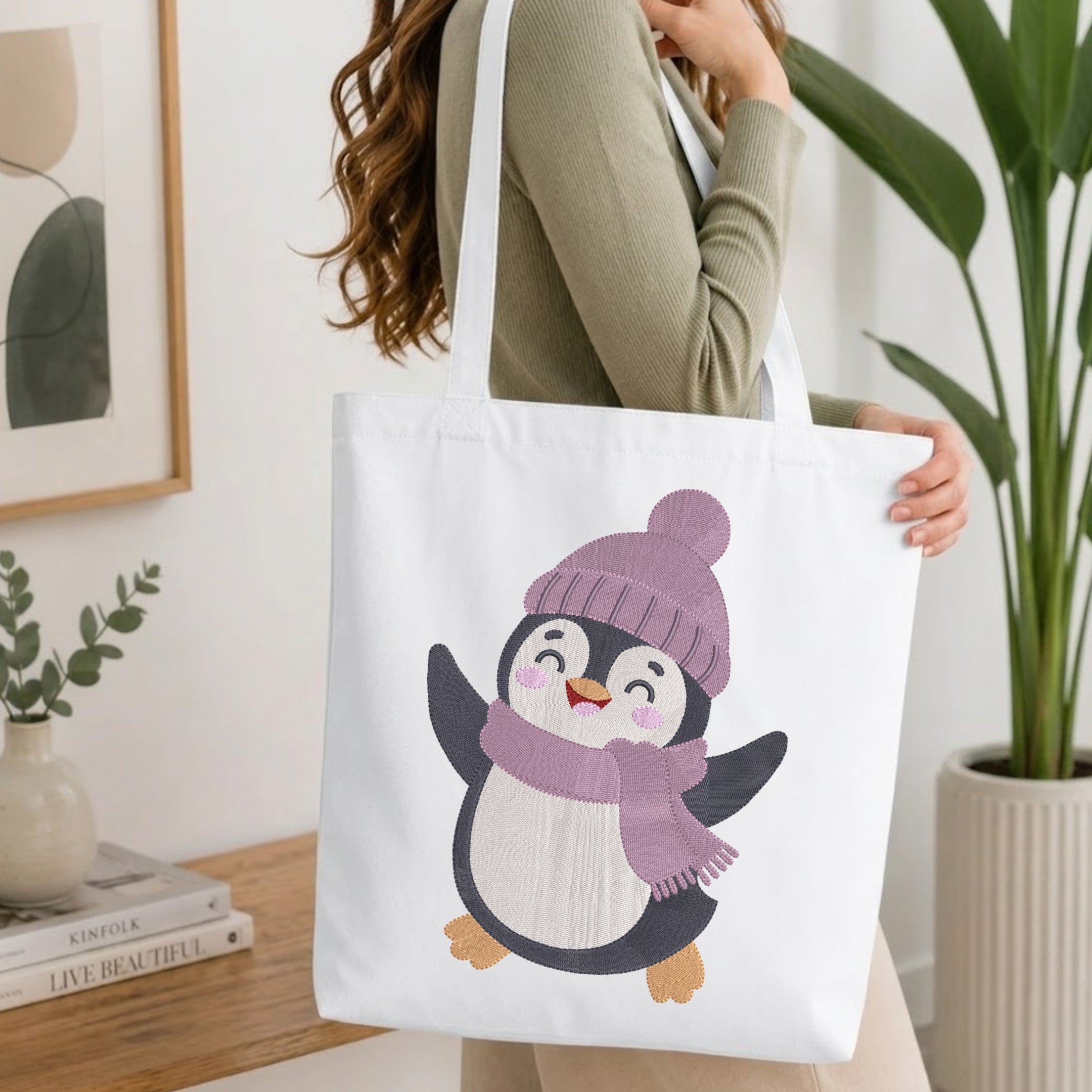 A woman wearing a sage green ribbed sweater carries a white canvas tote bag featuring a cute cartoon penguin wearing a purple knit hat and matching scarf.