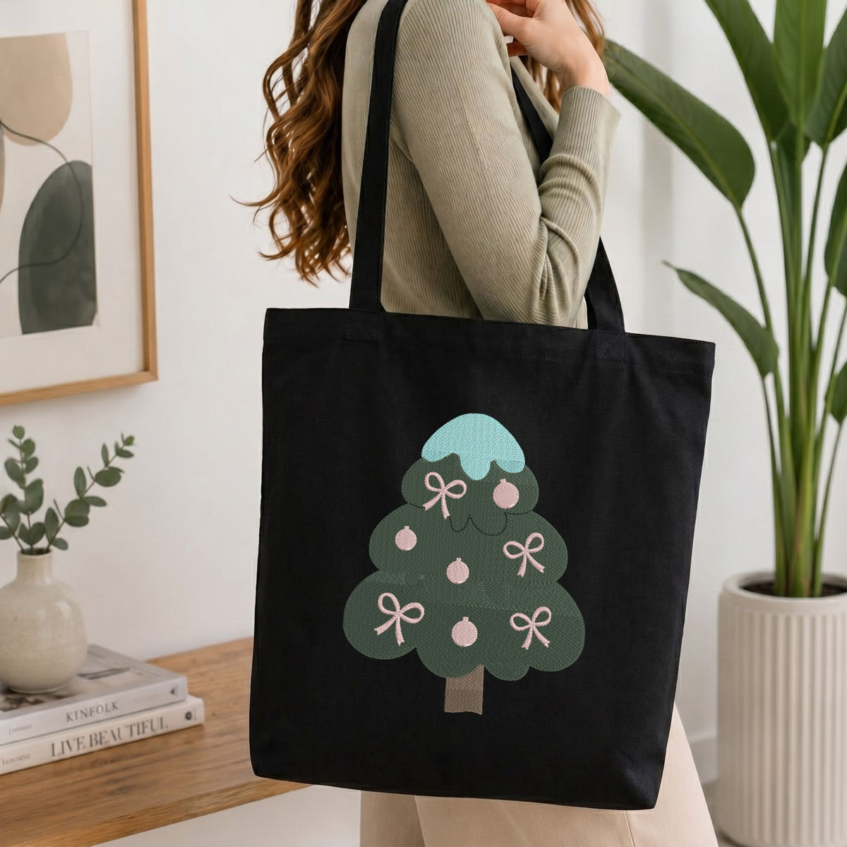 Woman carrying a black natural canvas tote bag featuring an embroidered Christmas tree design in a bright, modern interior setting.