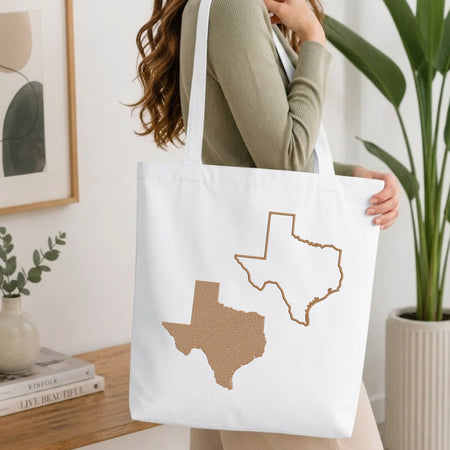 A woman in a sage green ribbed sweater carries a white canvas tote bag featuring a textured brown map of Texas and a simple brown outline of Texas. The background is a bright, modern room with a wooden table, framed art, and a potted plant.