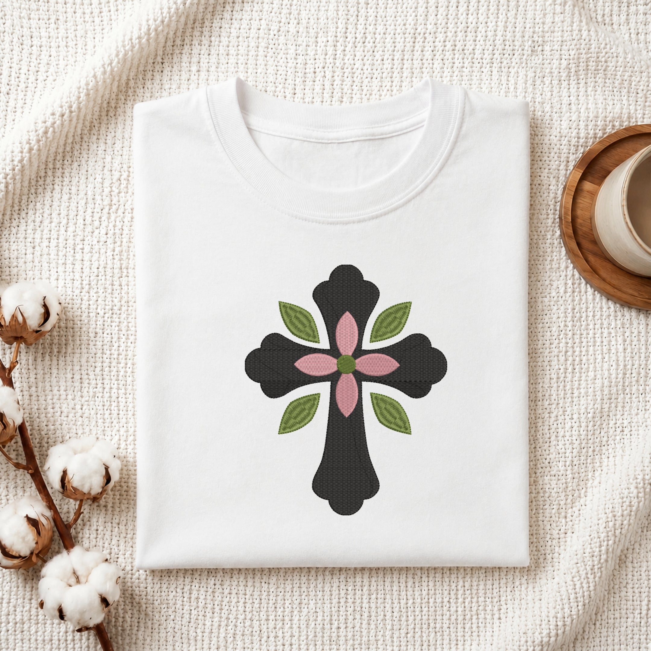 A neatly folded white crewneck t-shirt featuring a black floral cross embroidery design with pink and green accents, styled on a textured knit blanket next to cotton stems and a cup.