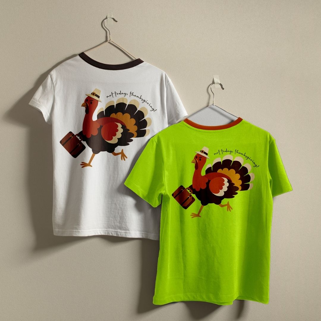 Two ringer t-shirts with a Thanksgiving turkey graphic hanging on metal hangers against a beige wall. The left shirt is white with a dark brown collar and features a large graphic of a walking turkey with a suitcase on the back.