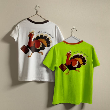 Two ringer t-shirts with a Thanksgiving turkey graphic hanging on metal hangers against a beige wall. The left shirt is white with a dark brown collar and features a large graphic of a walking turkey with a suitcase on the back.