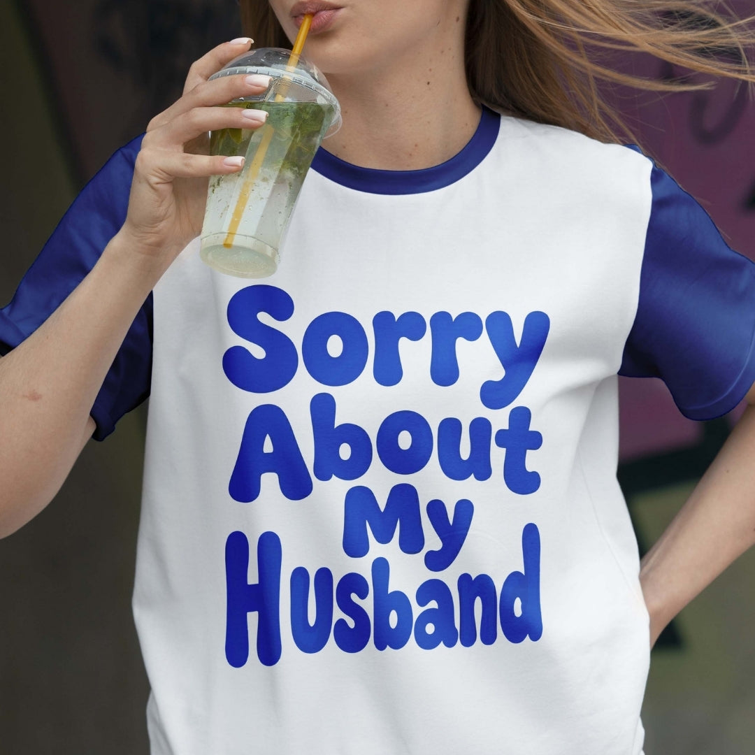 Person wearing a white t-shirt with blue text 'Sorry About My Husband' while holding a drink.