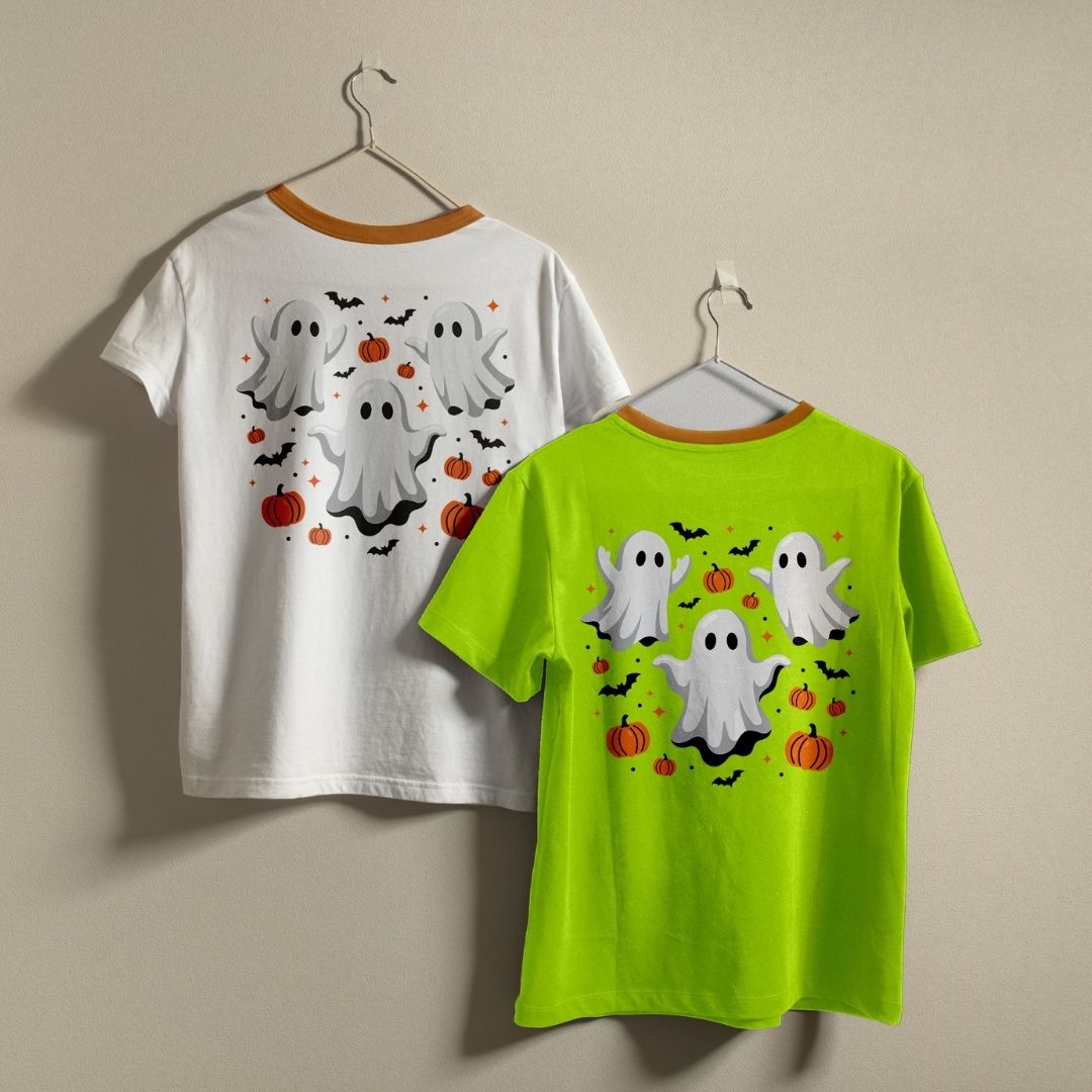 Two t-shirts hanging on a wall—one white and one bright green—both displaying the cute Halloween ghosts and pumpkins graphic print design.