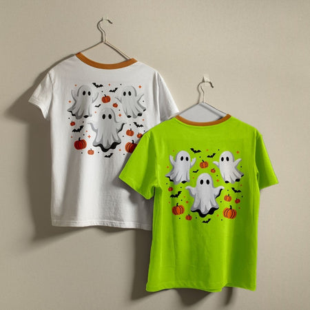 Two t-shirts hanging on a wall—one white and one bright green—both displaying the cute Halloween ghosts and pumpkins graphic print design.