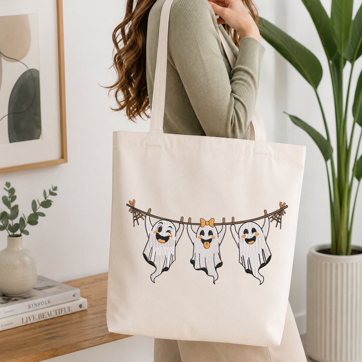 A woman wearing a sage green ribbed long-sleeve top carries a cream canvas tote bag with a graphic of three cute ghosts hanging from a clothesline. The setting is a bright, modern room with a wooden table, books, a small plant, and a large green potted plant in the background.