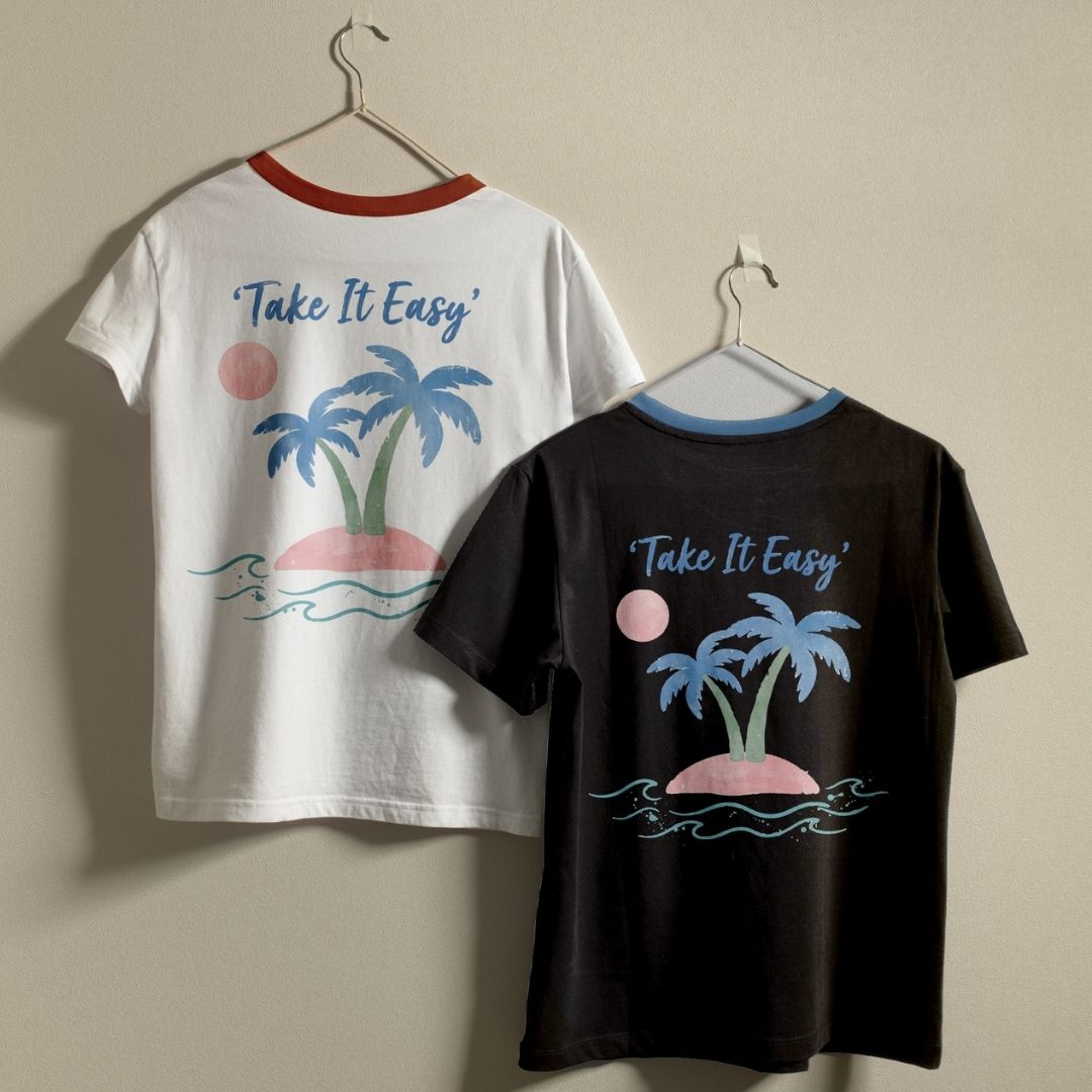 Two ringer t-shirts hanging on hangers against a plain beige wall. The left shirt is white with a red collar trim and features a tropical graphic with blue palm trees, a pink island, and the text 'Take It Easy'.