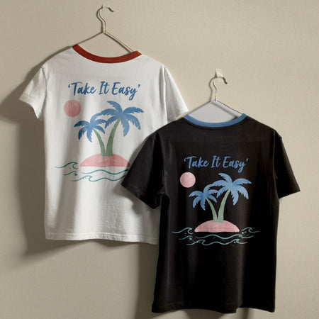 Two ringer t-shirts hanging on hangers against a plain beige wall. The left shirt is white with a red collar trim and features a tropical graphic with blue palm trees, a pink island, and the text 'Take It Easy'.