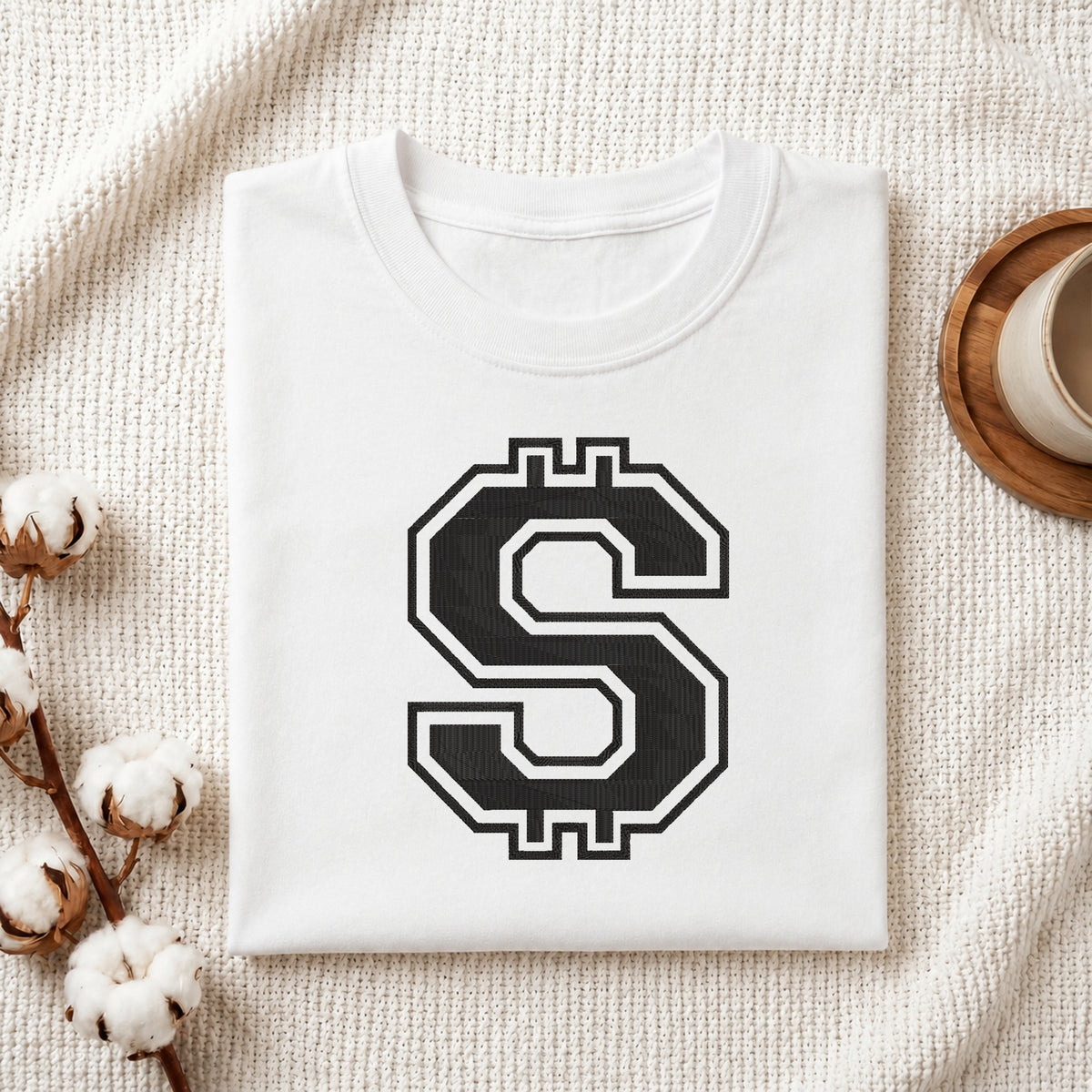 A folded white crewneck t-shirt with a large black and white stylized 'S' graphic printed on the chest, styled on a textured cream knit blanket with cotton bolls and a wooden cup in the background.