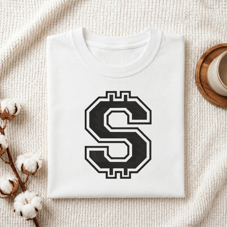 A folded white crewneck t-shirt with a large black and white stylized 'S' graphic printed on the chest, styled on a textured cream knit blanket with cotton bolls and a wooden cup in the background.