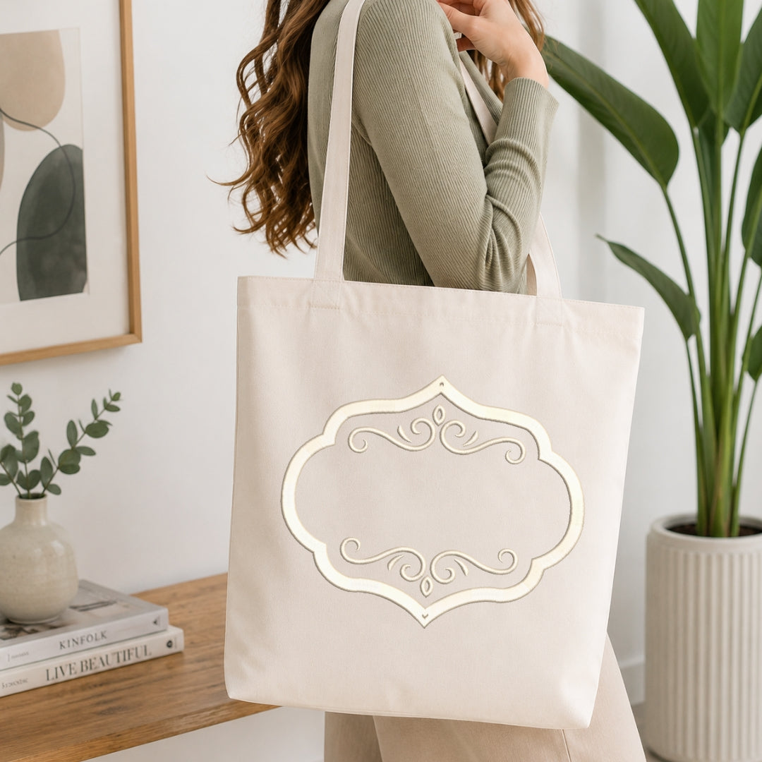 A woman wearing a sage green ribbed sweater carries a large cream canvas tote bag with a decorative white scalloped label outline on the front, set in a bright, modern room with a wooden table, framed art, and a potted plant.