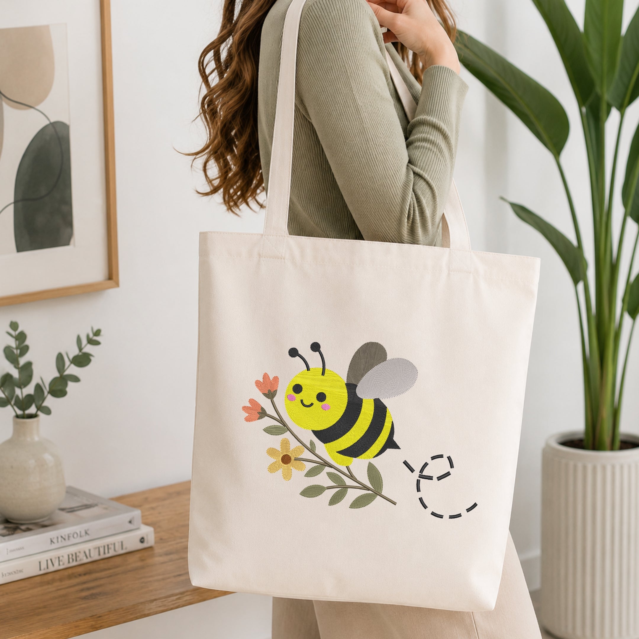 A woman wearing a sage green ribbed sweater carries a cream canvas tote bag with a cute yellow and black bumblebee graphic on the front, set in a bright, modern room with a wooden table and potted plants.