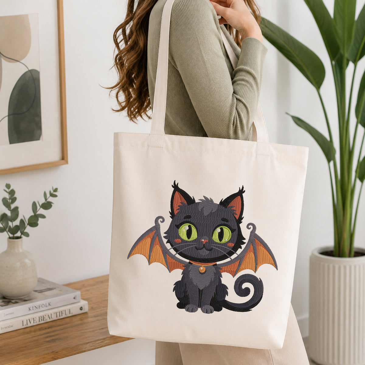 A woman wearing a sage green ribbed sweater carries a cream canvas tote bag with a cute cartoon bat cat graphic. The tote bag features a black cat with orange wings and green eyes.
