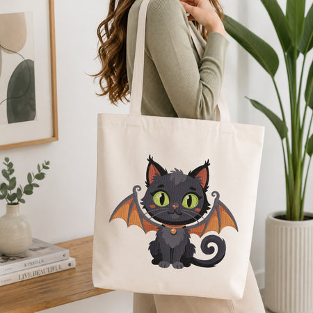 A woman wearing a sage green ribbed sweater carries a cream canvas tote bag with a cute cartoon bat cat graphic. The tote bag features a black cat with orange wings and green eyes.