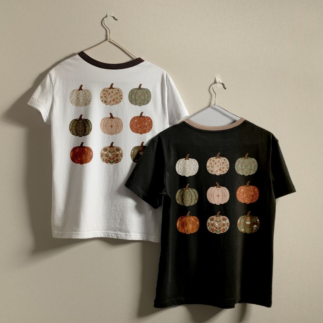 Two children's short-sleeve ringer T-shirts hanging on hangers against a plain beige wall. The left shirt is white with a dark brown collar and back graphic of 12 pumpkins in muted autumn colors.
