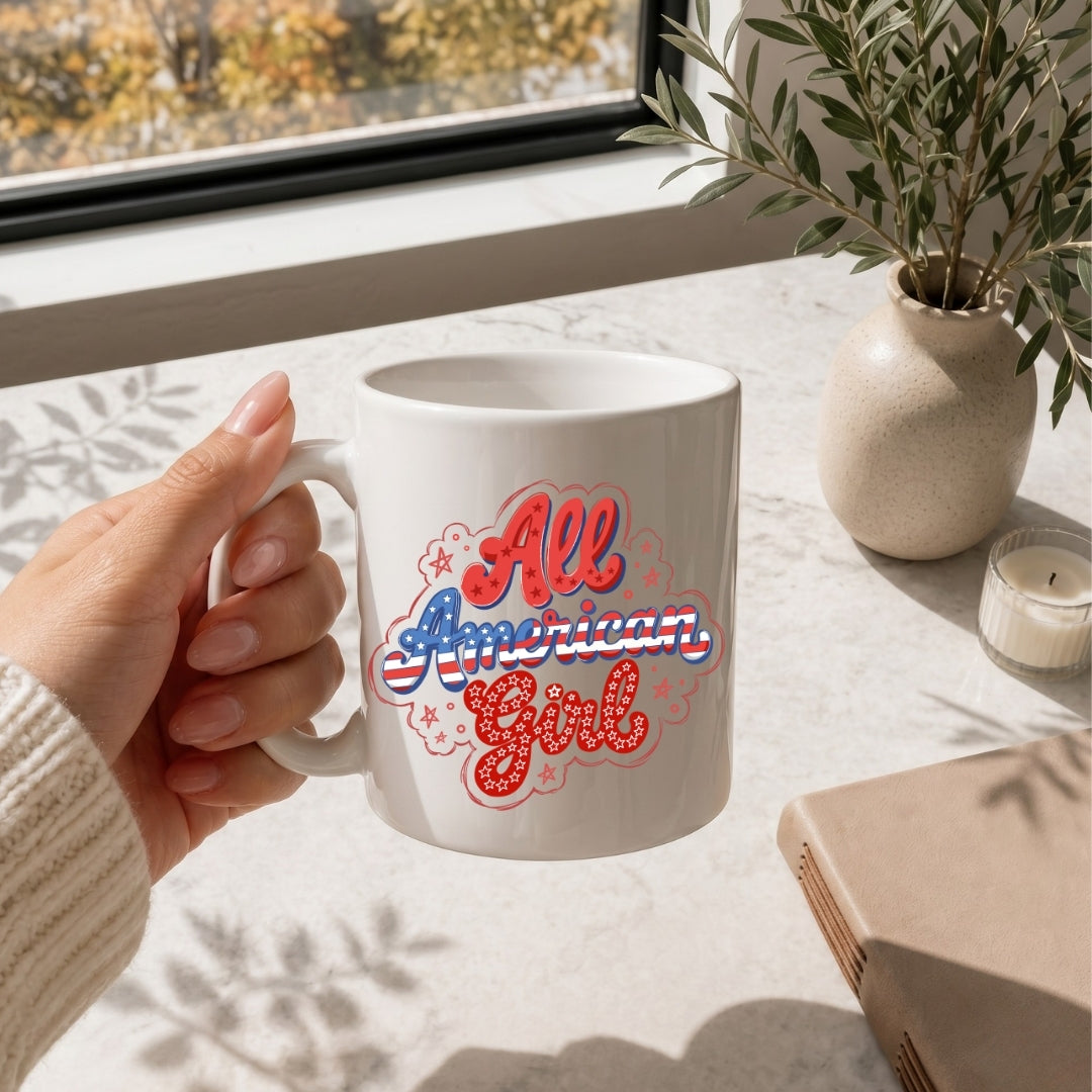 White mug with 'All American Girl' design held by a hand on a table with a window and plant in the background.
