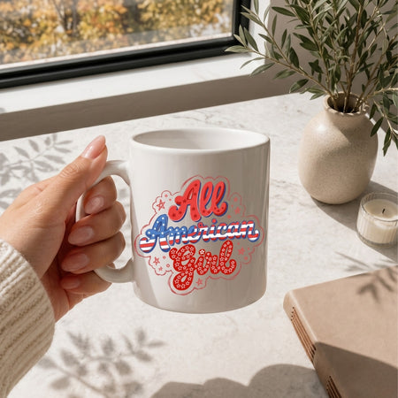 White mug with 'All American Girl' design held by a hand on a table with a window and plant in the background.