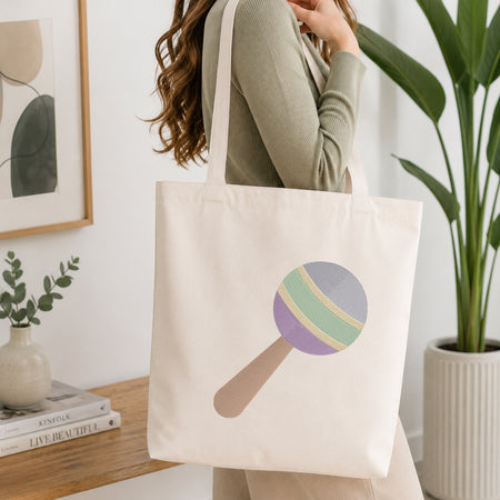 A woman wearing a sage green ribbed sweater carries a cream canvas tote bag with a colorful lollipop graphic. The background is a bright, modern room with a wooden shelf, framed art, and a potted plant.