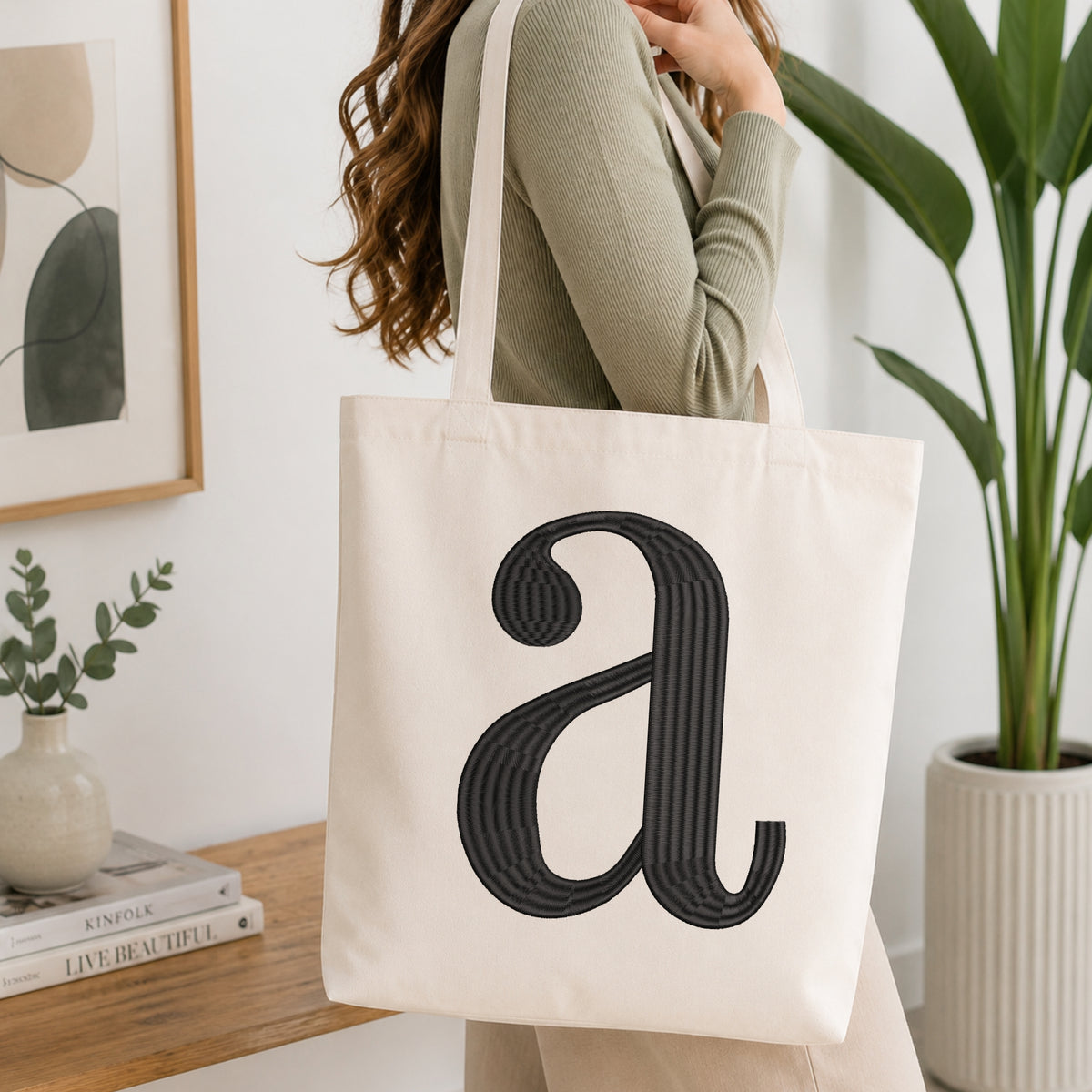 A woman wearing a sage green ribbed long-sleeve top carries a cream canvas tote bag with a large black textured letter 'a' on the front, set in a bright, modern room with a wooden console table, framed art, and a potted plant.