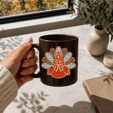 A hand holds a dark brown ceramic mug featuring a stylized orange and patterned turkey design, set on a light stone surface near a window with a plant and candle in the background.
