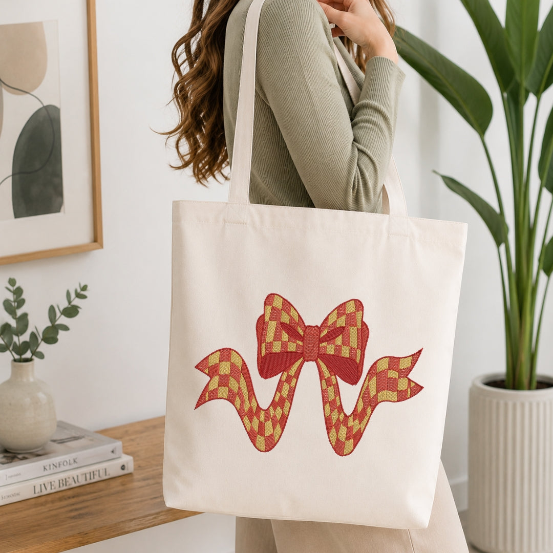 A person wearing a ribbed sage green long-sleeve top carries a cream canvas tote bag with a red and gold checkered bow graphic on the front, set in a bright, modern room with a wooden table and potted plants.