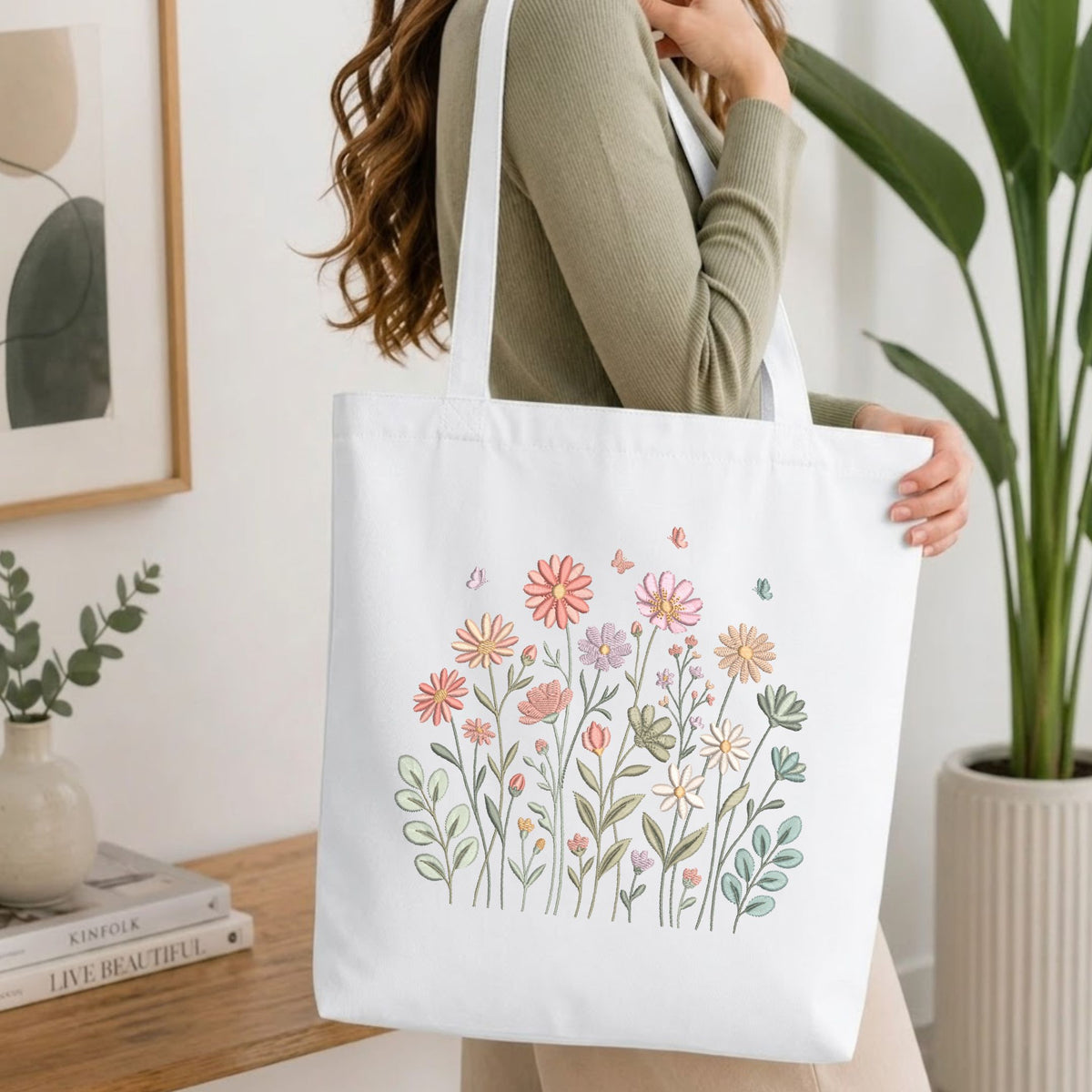 A woman wearing a sage green ribbed long-sleeve top carries a white canvas tote bag decorated with a colorful hand-drawn floral and butterfly pattern. The setting is a bright, modern room with a wooden table, books, a framed print, and a potted green plant.