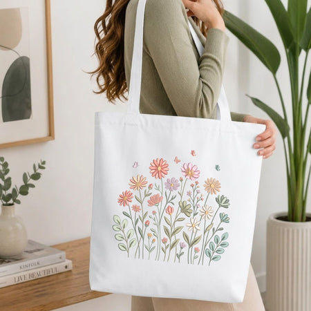 A woman wearing a sage green ribbed long-sleeve top carries a white canvas tote bag decorated with a colorful hand-drawn floral and butterfly pattern. The setting is a bright, modern room with a wooden table, books, a framed print, and a potted green plant.