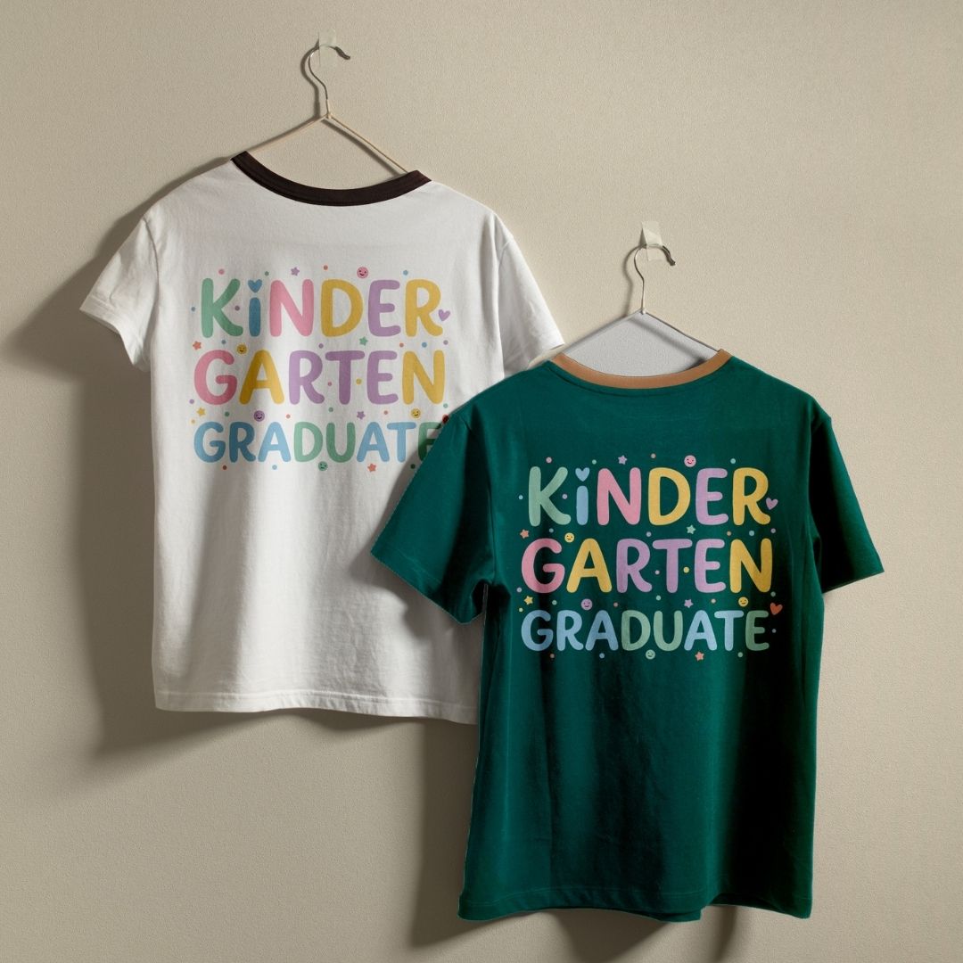 Two children's ringer t-shirts hanging on metal hangers against a plain beige wall. The back of each shirt features the text 'KINDER GARTEN GRADUATE' in a colorful retro font with small star and dot accents.