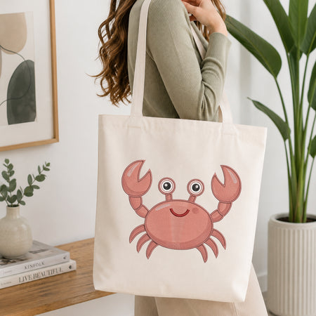 A cream canvas tote bag with a pink cartoon crab graphic, carried by a woman in a sage green top in a bright, modern room with a potted plant and books.