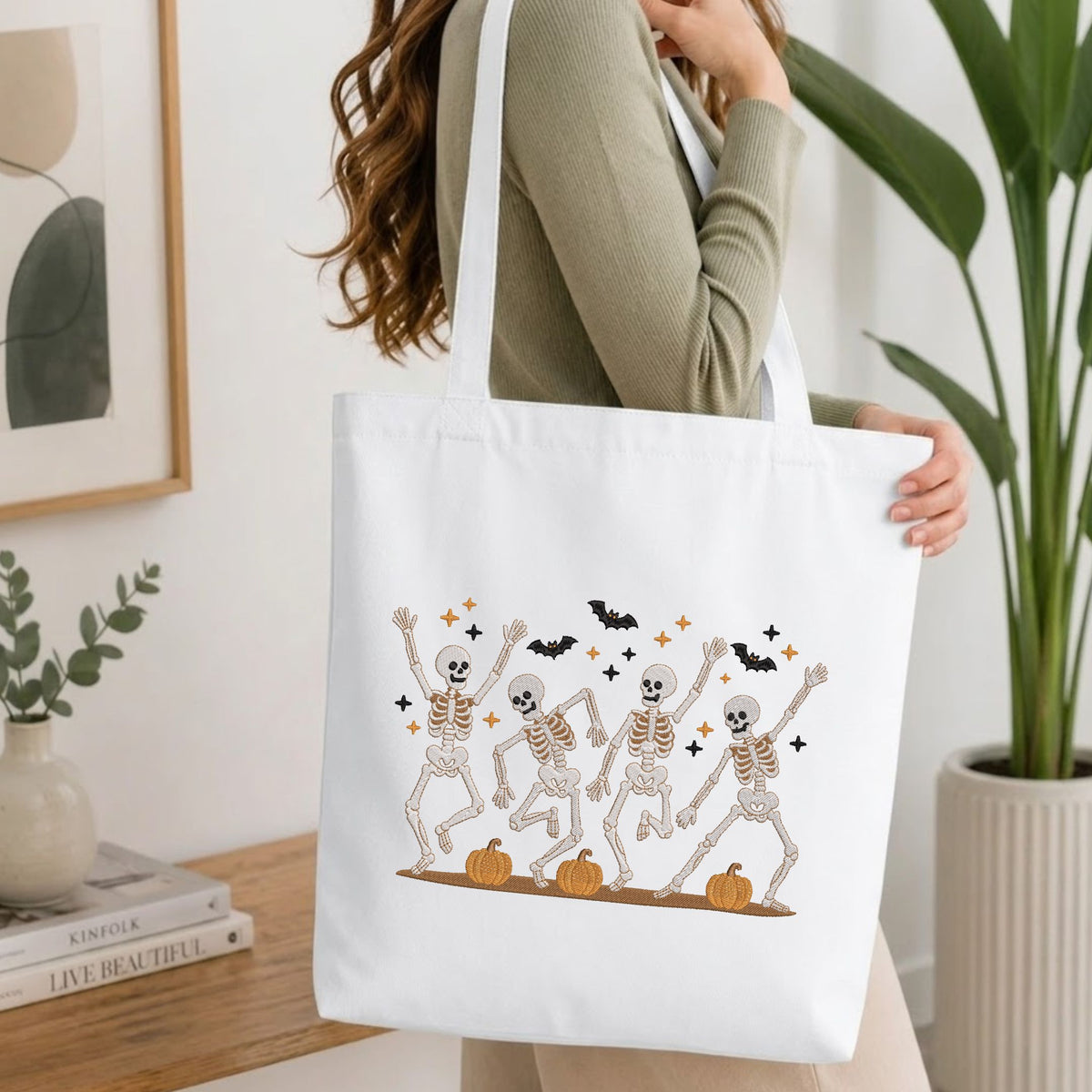 Woman carrying a white natural canvas tote bag featuring an embroidered dancing skeletons Halloween design in a bright, modern interior setting   