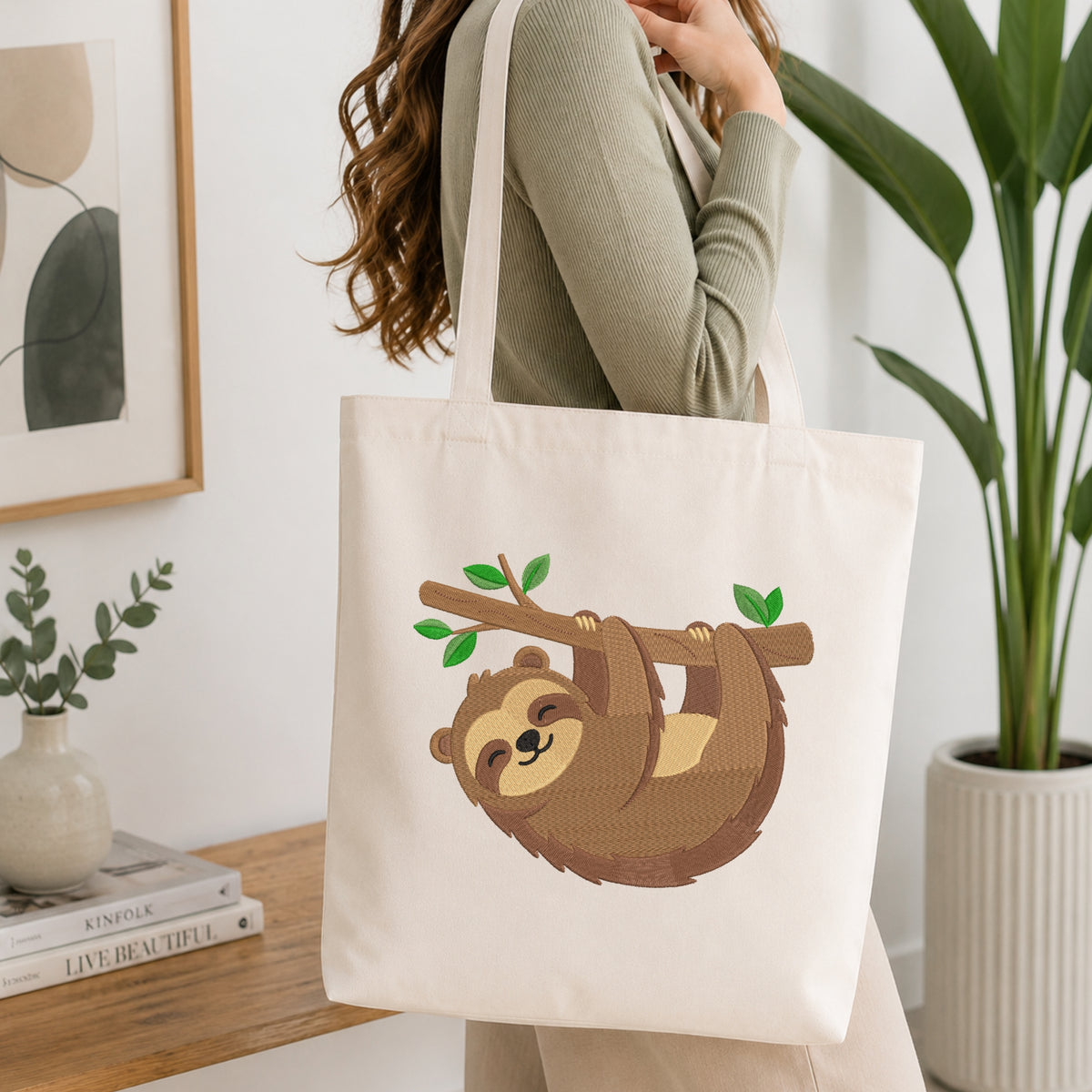 A woman wearing a sage green ribbed sweater carries a cream canvas tote bag with a brown sloth graphic hanging from a branch. The scene is set in a bright, modern room with a wooden table, books, a small plant, and a large green potted plant in the background.