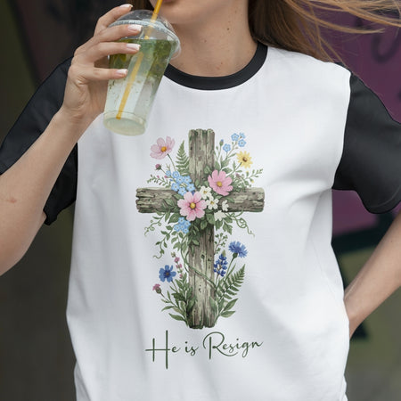Person wearing a white t-shirt with a floral cross design and 'He is Risen' text, holding a drink.