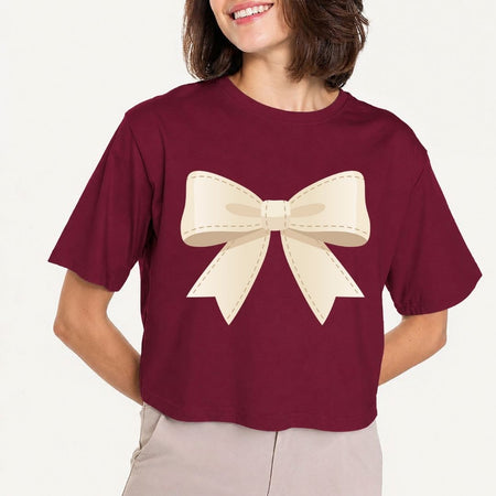 Front view mockup of a woman wearing a maroon cropped t-shirt displaying a large, off-white cream bow graphic with stitched detail edging.