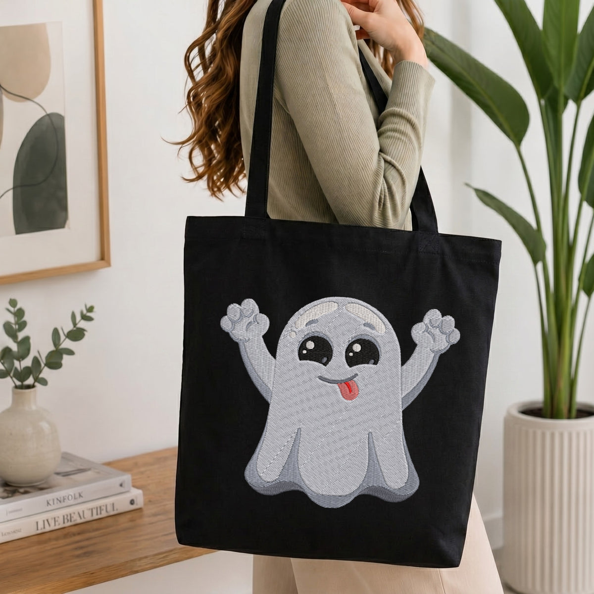 A woman wearing a beige ribbed sweater carries a black canvas tote bag with a large embroidered cute ghost design on the front, set in a bright, modern room with a wooden table, books, and a potted plant.