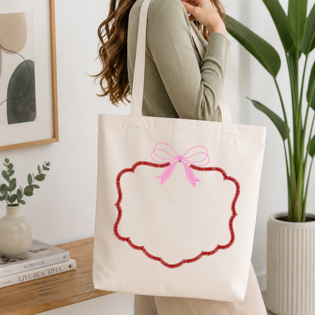 A woman wearing a sage green ribbed sweater carries a cream canvas tote bag with a pink bow and red scalloped border graphic, set in a bright, modern room with a wooden table, books, and a potted plant.