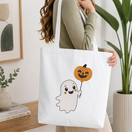 Woman carrying a white natural canvas tote bag featuring a cute ghost machine embroidery design in a bright, modern interior setting