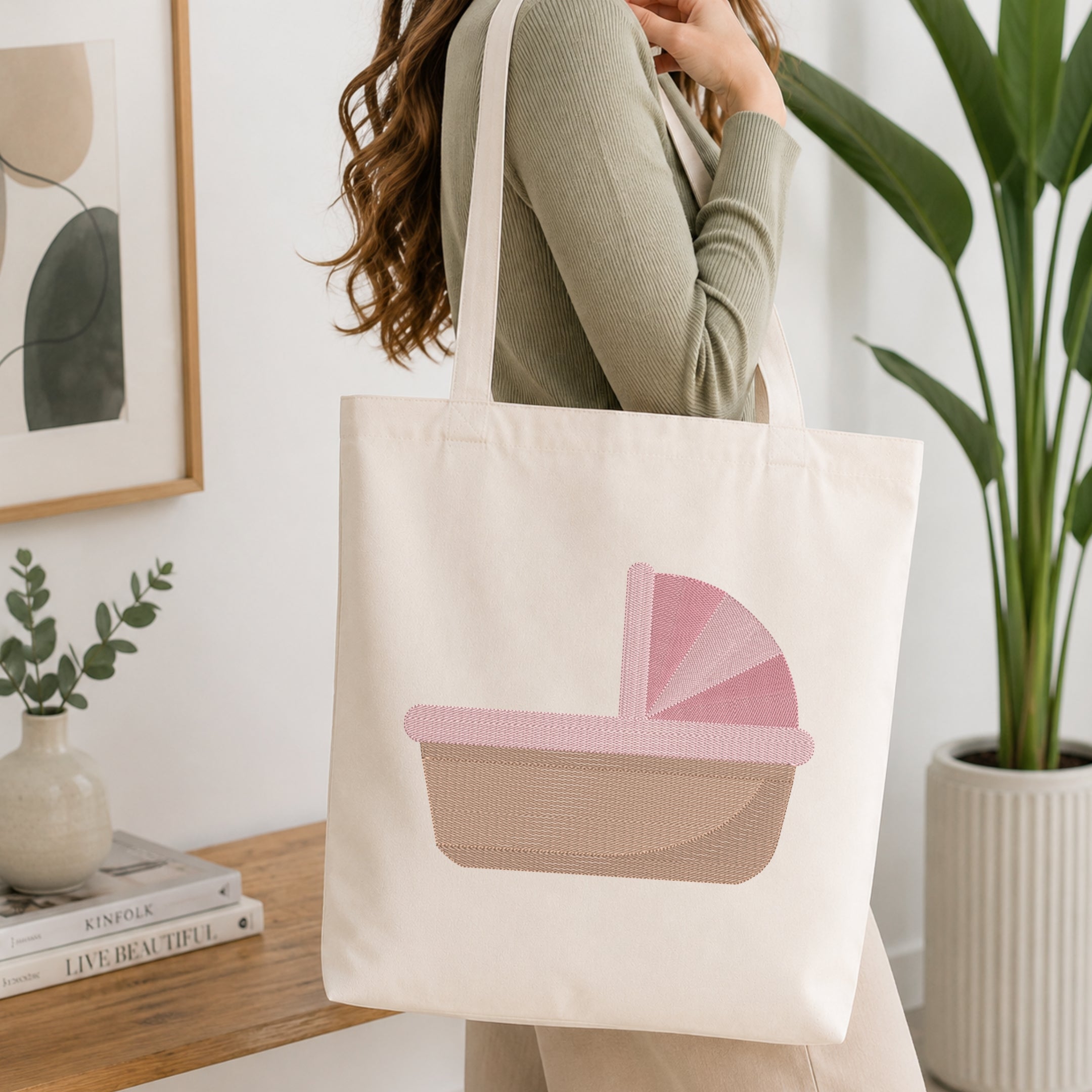 A woman wearing a sage green ribbed long-sleeve top carries a cream canvas tote bag with a pink and beige baby stroller graphic, set in a bright, modern room with a wooden table, framed art, and a potted plant.