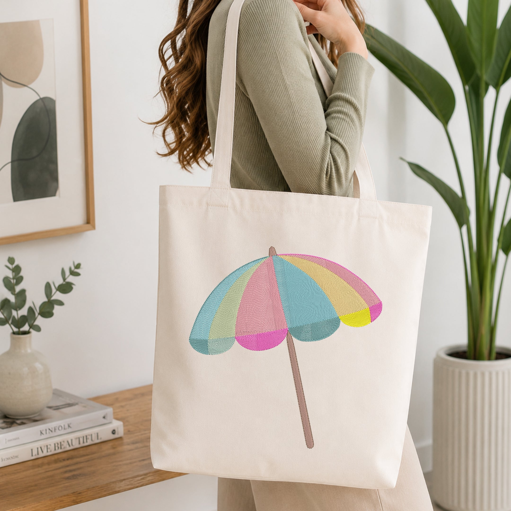 A woman wearing a sage green ribbed sweater carries a cream canvas tote bag with a colorful pastel beach umbrella graphic. The setting is a bright, modern room with a wooden console table, a potted green plant, and a framed abstract print.