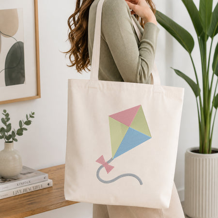 A person holding a natural canvas tote bag printed with an embroidered colorful kite design, set against a bright interior room background with green plants.