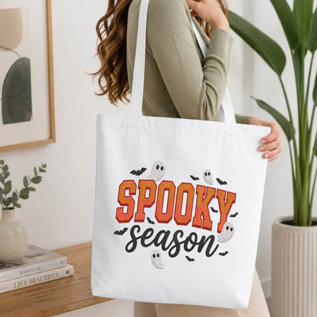 Woman carrying a white natural canvas tote bag featuring an embroidered spooky season ghost design in a bright, modern interior setting.   