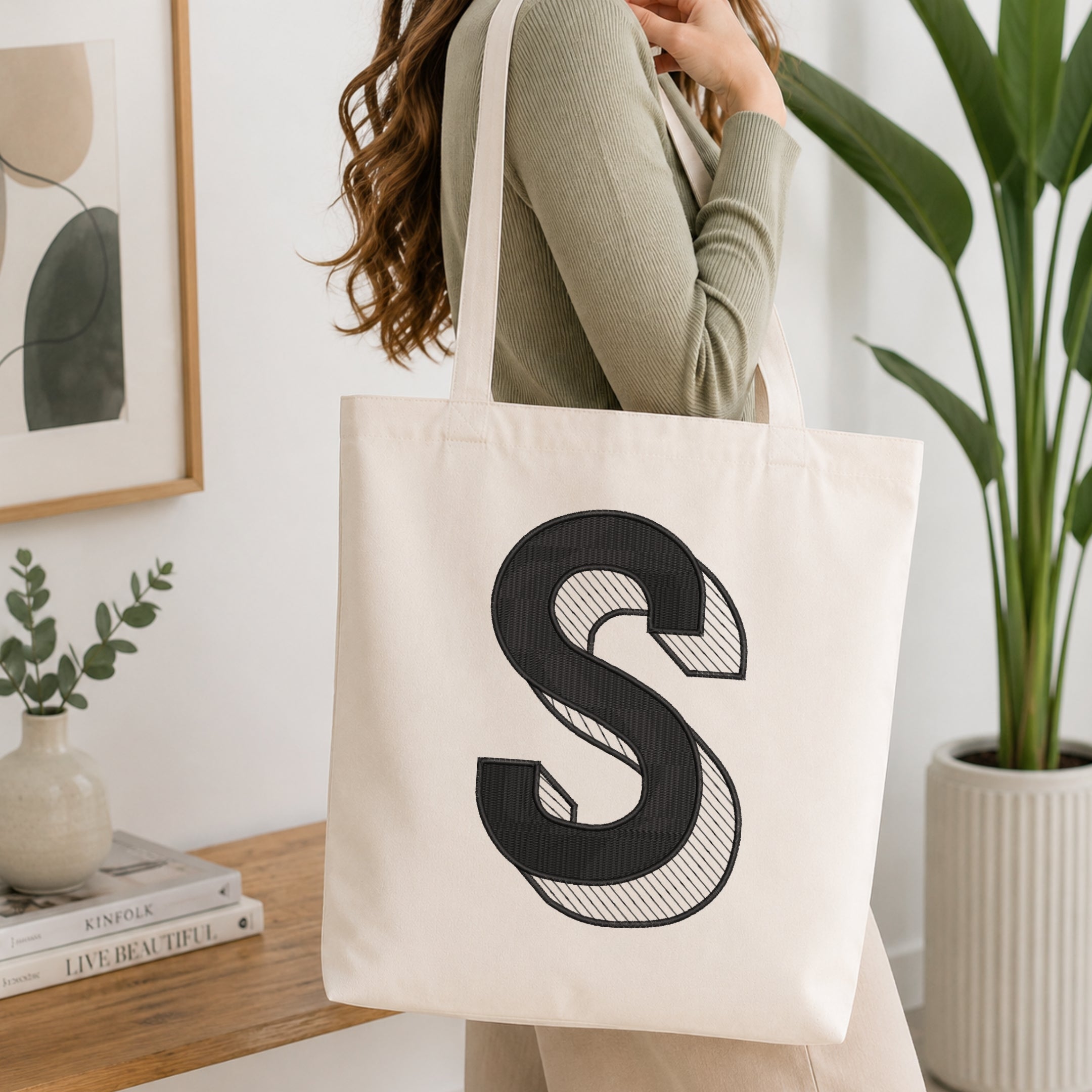 A woman wearing a sage green ribbed long-sleeve top carries a large off-white canvas tote bag with a bold black embroidered letter 'S' on the front, set in a bright, modern room with a wooden table, framed art, and a potted plant.