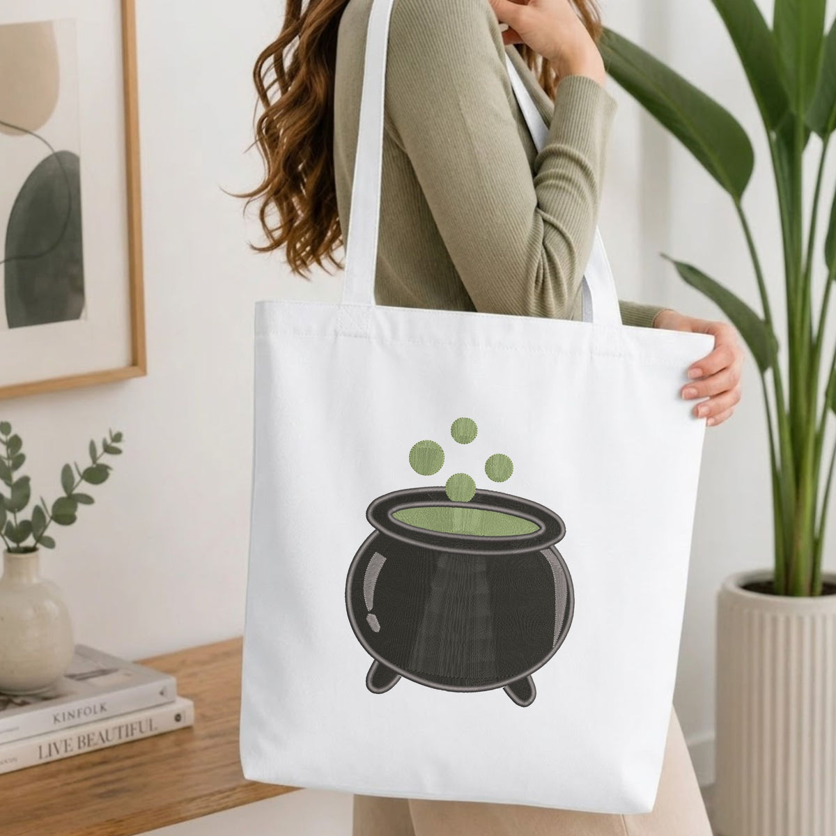 Woman carrying a white natural canvas tote bag featuring an embroidered witch cauldron design in a bright, modern interior setting.