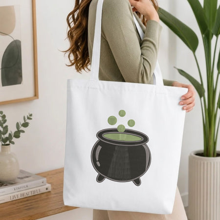 Woman carrying a white natural canvas tote bag featuring an embroidered witch cauldron design in a bright, modern interior setting.