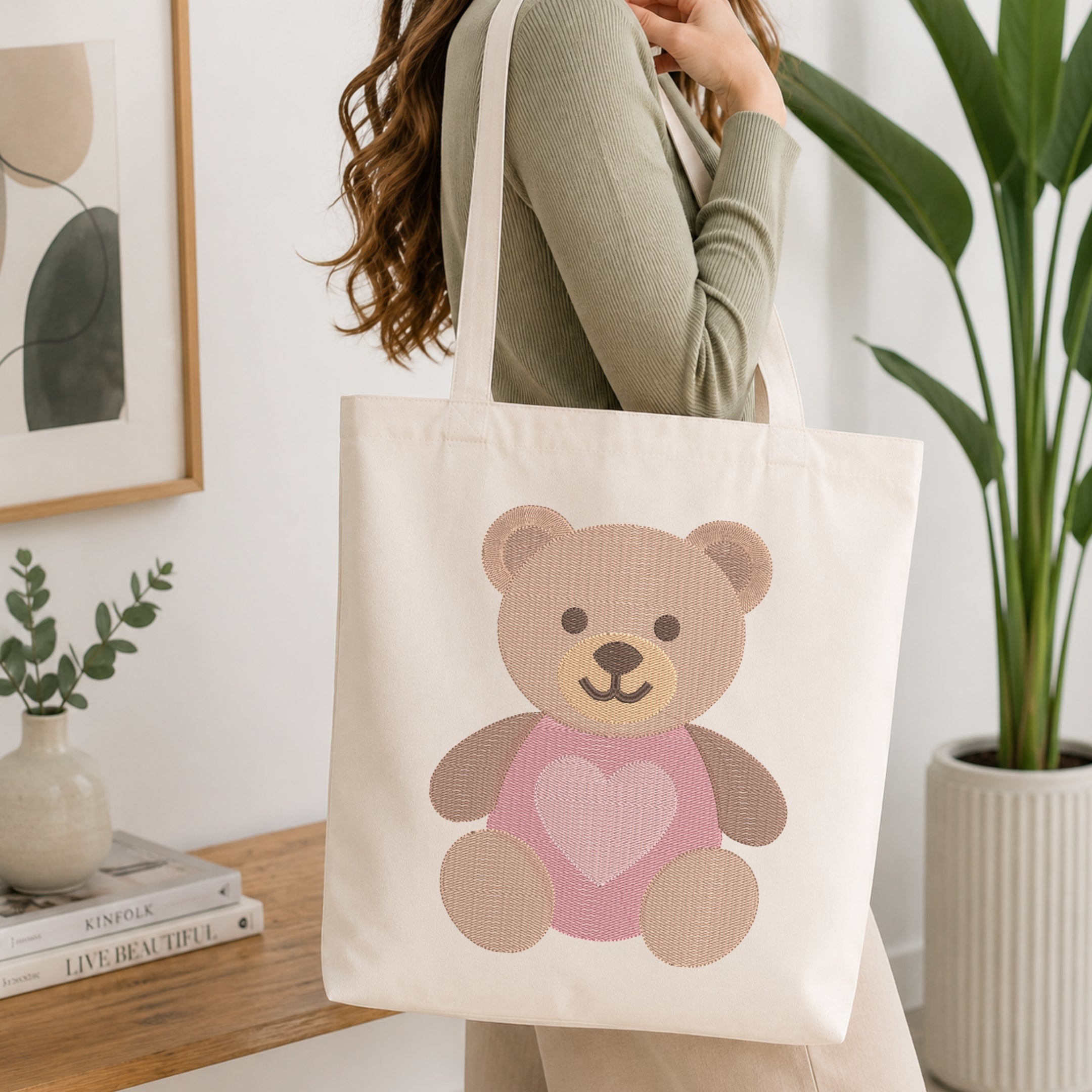 A woman wearing a sage green ribbed sweater carries a cream canvas tote bag with a teddy bear graphic. The bear is tan with a pink heart on its chest.