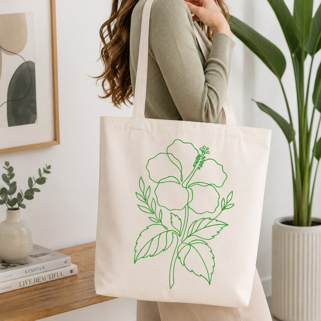 A person carrying a cream-colored canvas tote bag that is decorated with the same green, line-art hibiscus embroidery design.