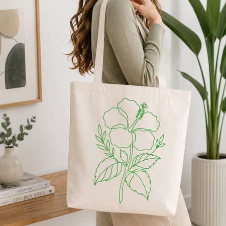 A person carrying a cream-colored canvas tote bag that is decorated with the same green, line-art hibiscus embroidery design.