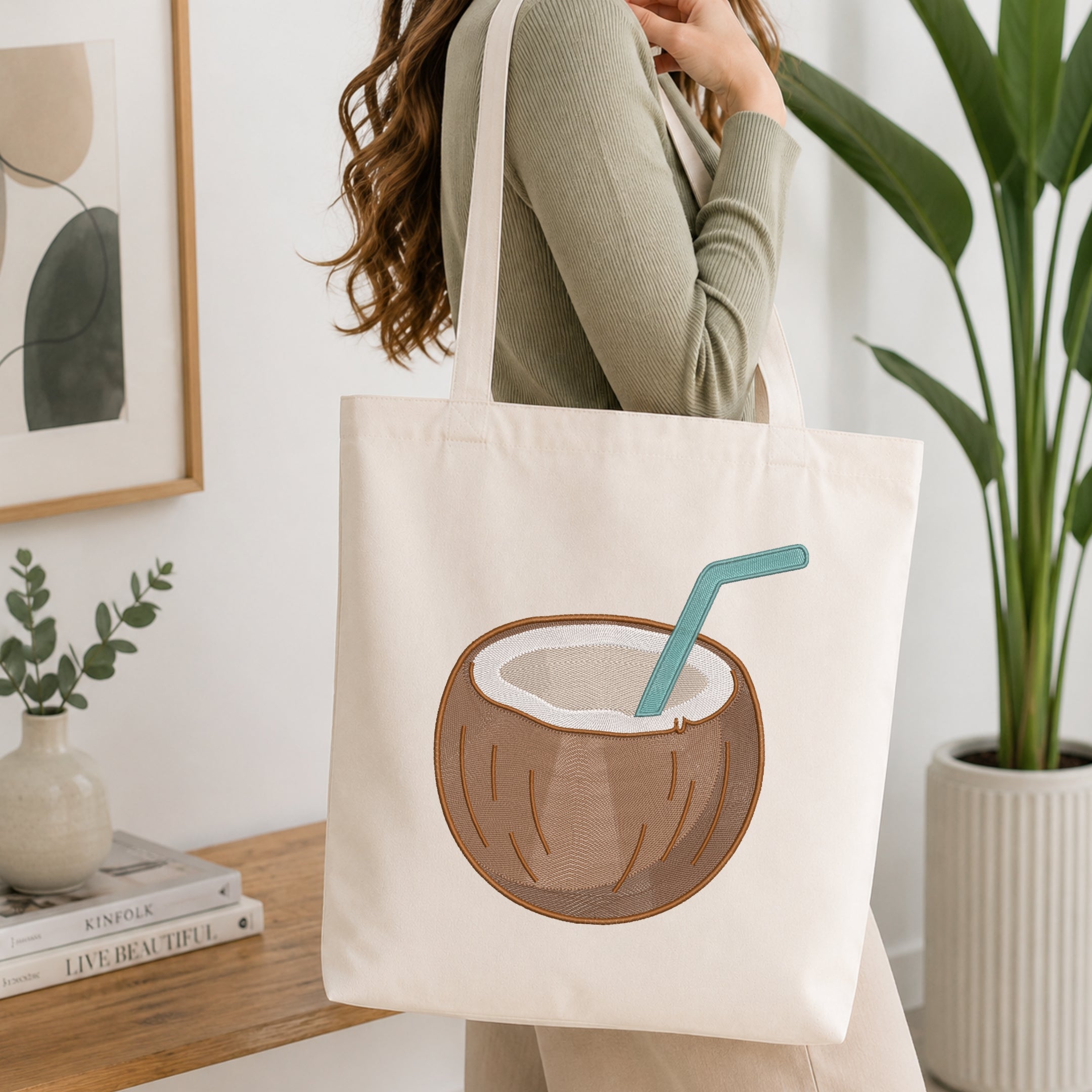 A woman wearing a sage green ribbed sweater carries a cream canvas tote bag with an embroidered coconut drink design featuring a teal straw, set in a bright, modern room with a wooden table, framed art, and a potted plant.