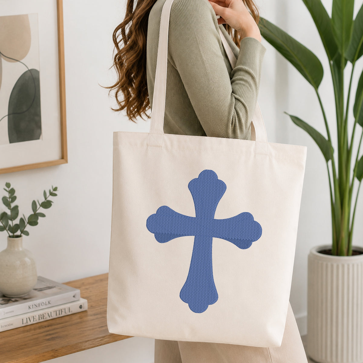 A woman wearing a sage green ribbed top carries a cream canvas tote bag with a large blue cross design on the front, set in a bright, modern room with a wooden table, books, and a potted plant.