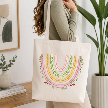 A woman wearing a sage green ribbed sweater carries a cream canvas tote bag with a colorful pastel rainbow design featuring pink, yellow, and green stripes with floral accents.